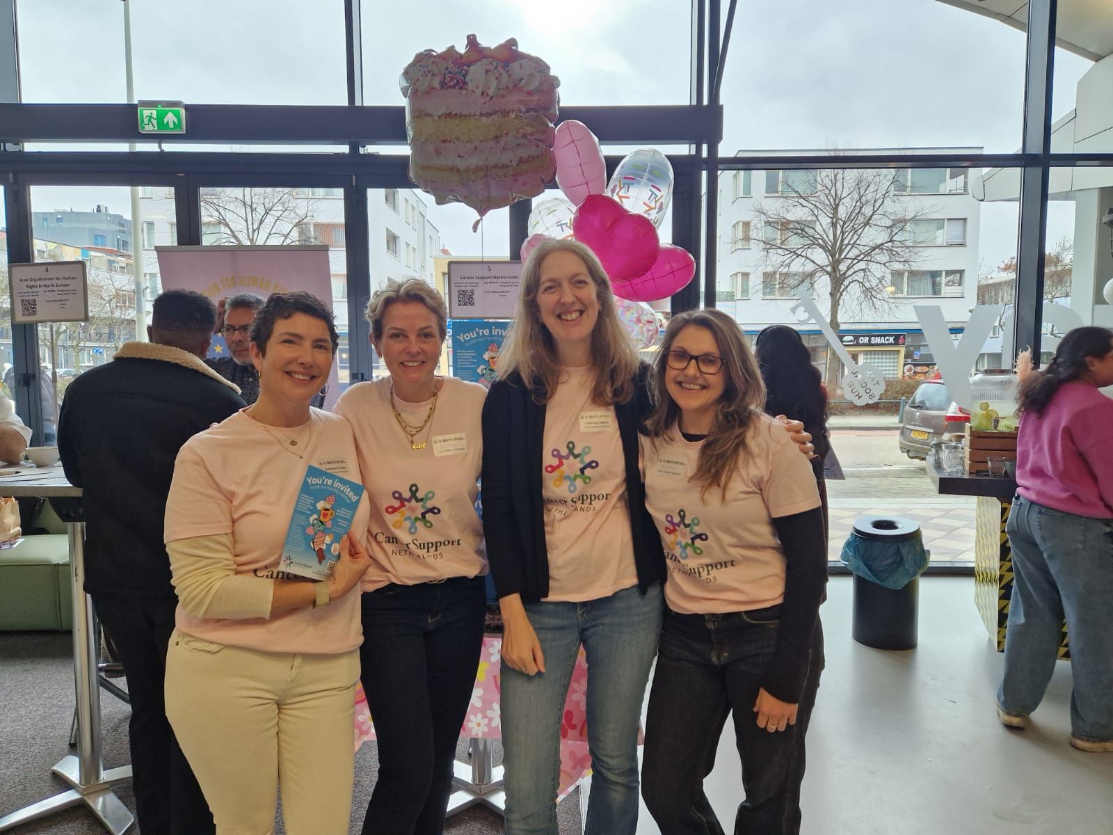 Four CSN volunteers in pink Cancer Support Netherlands T-shirts at an information stand with pink balloons