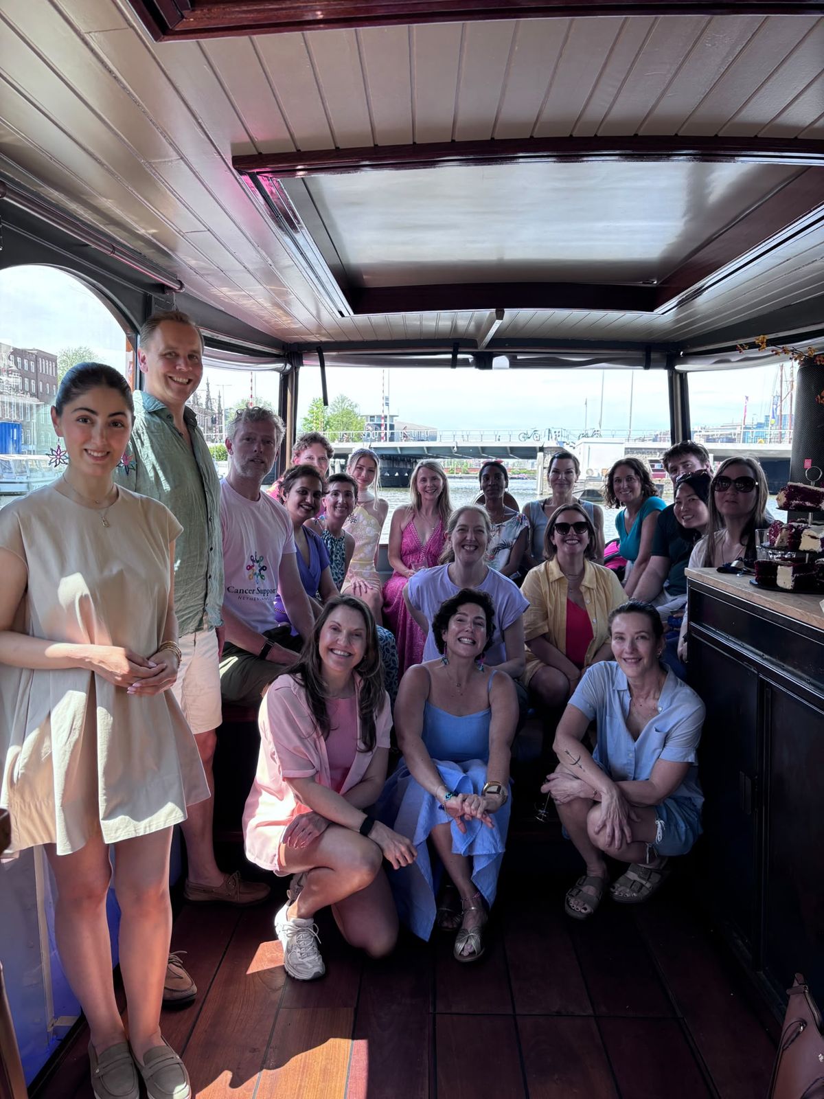 Around twenty Cancer Support Netherlands members smiling together inside a canal boat on a sunny day